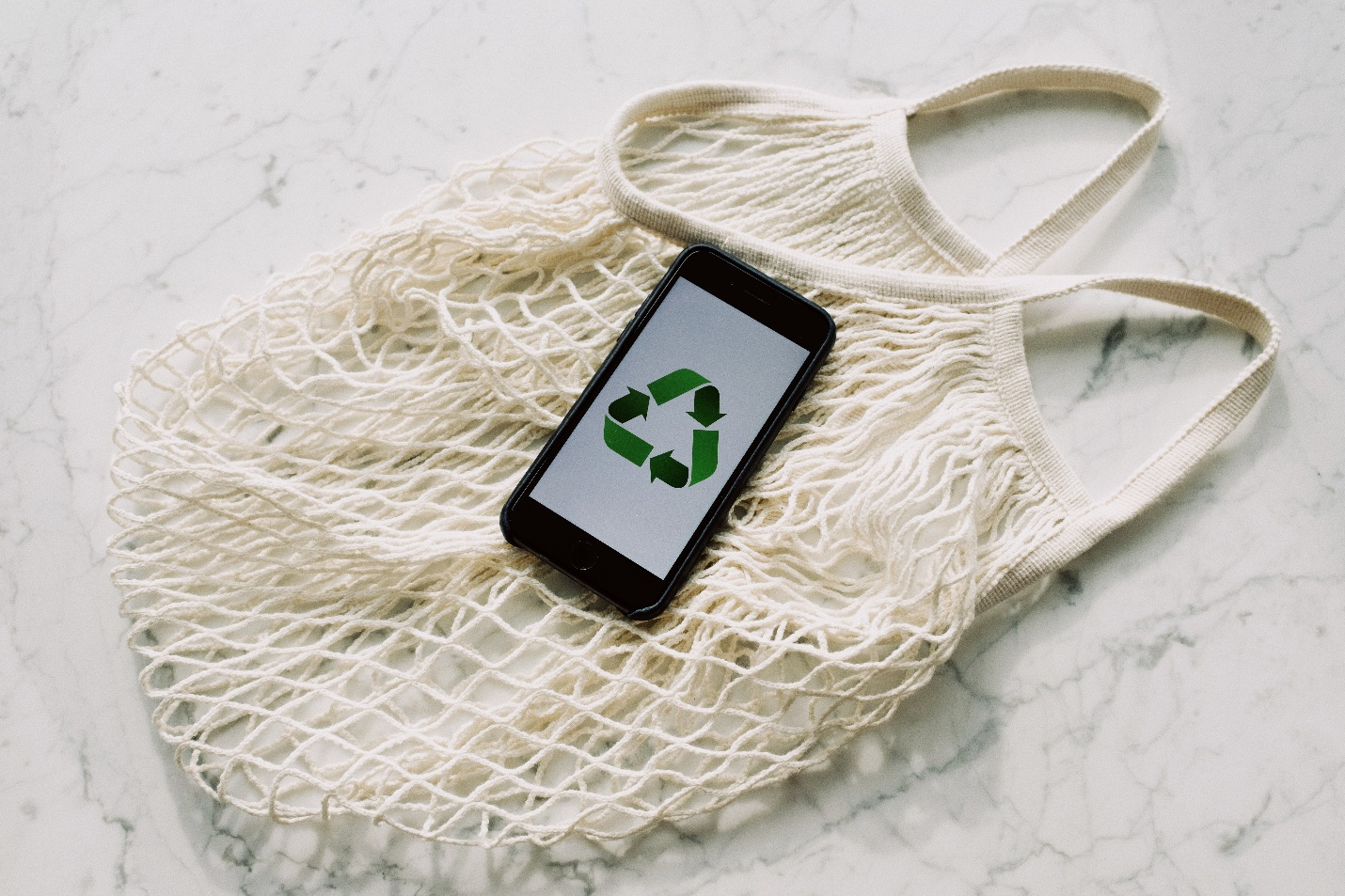 A phone showing a recycle sign placed on top of a white mesh bag.